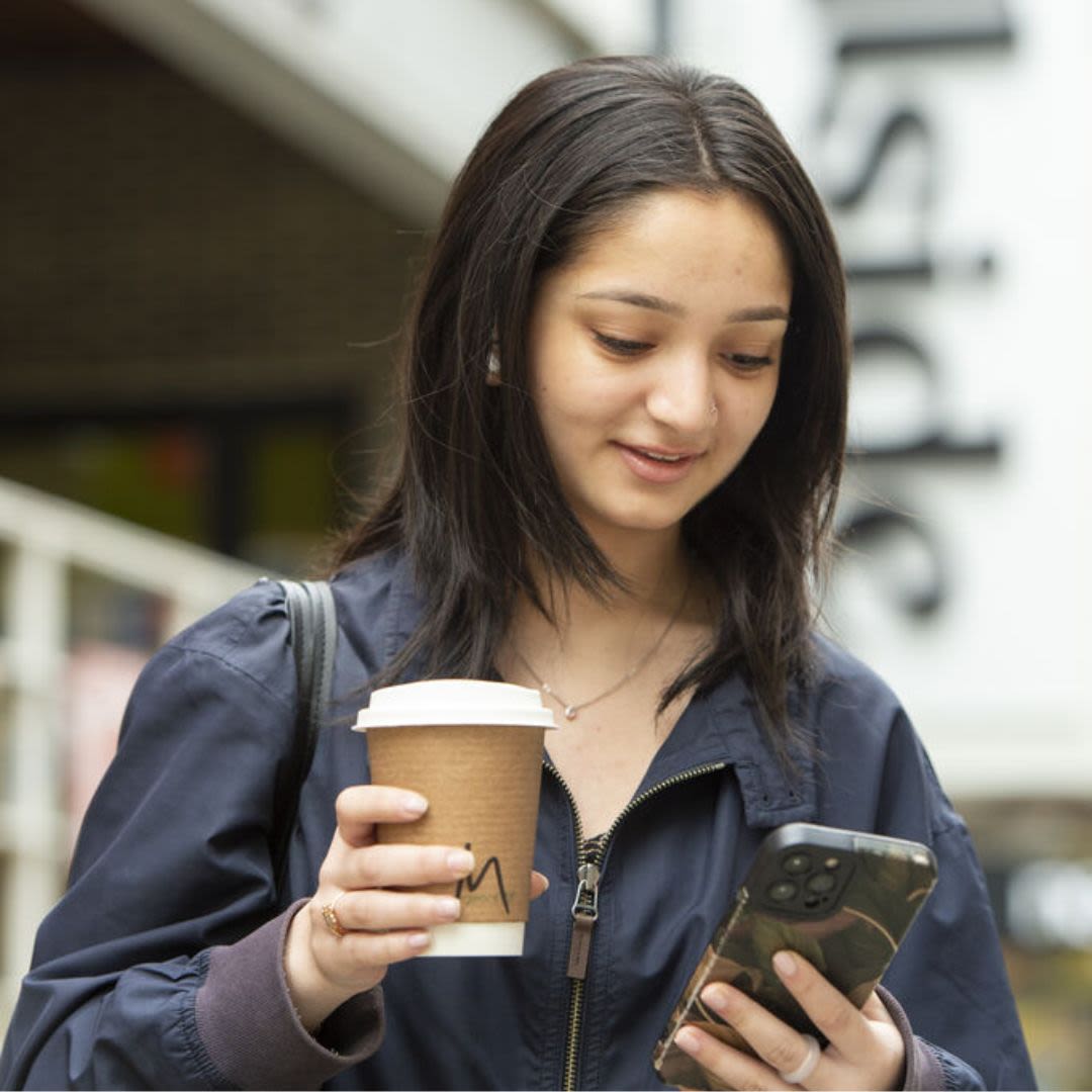 A student holds a takeaway coffee in one hand and her phone in the other outside Hillside Coffee