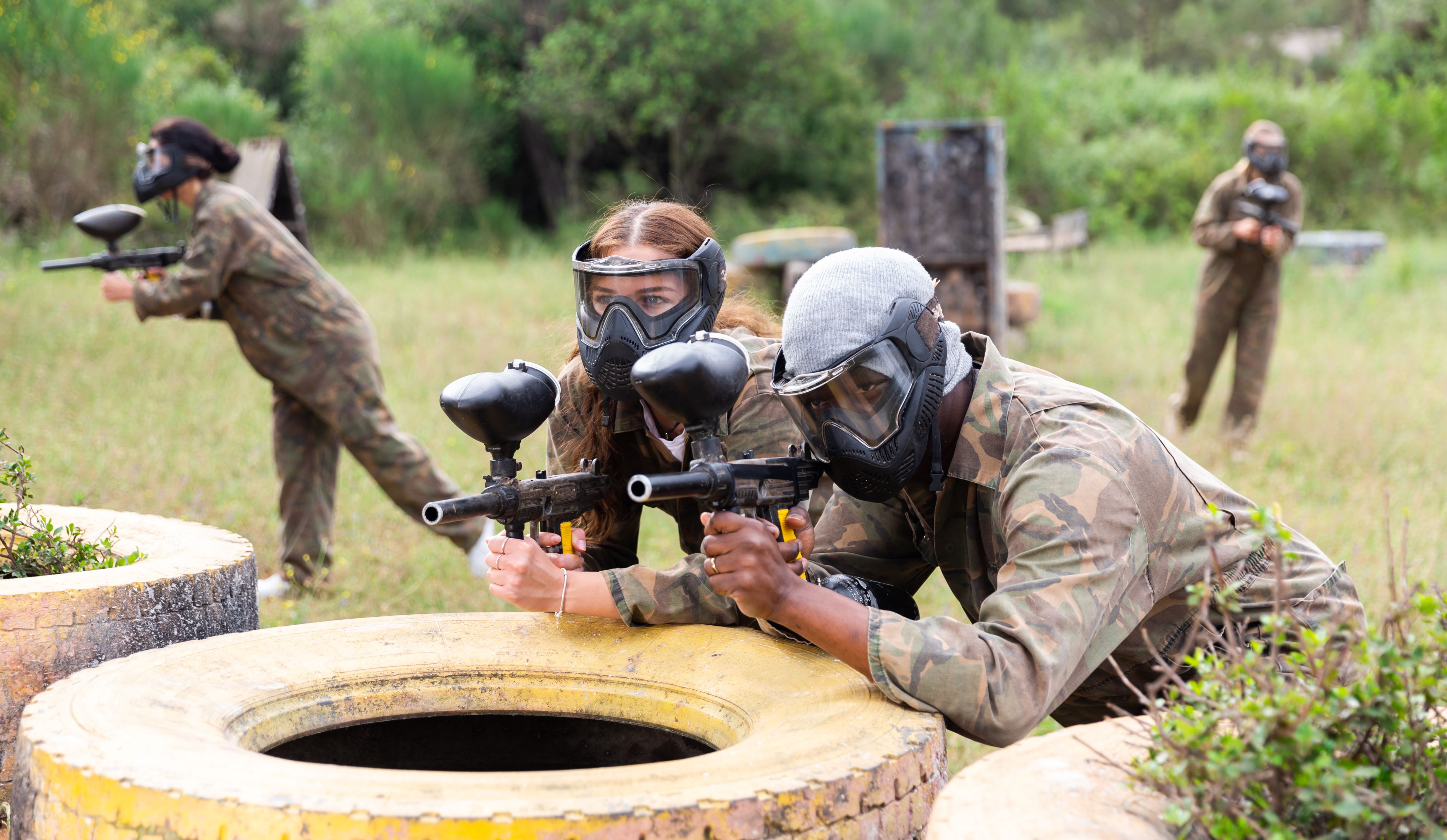 Two young people with paintball guns leaning on a tire in a field 