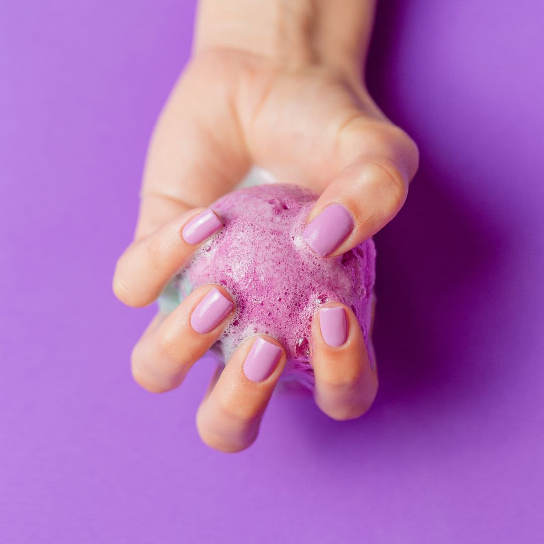 A hand with purple nail polish holds a fizzing purple bath bomb