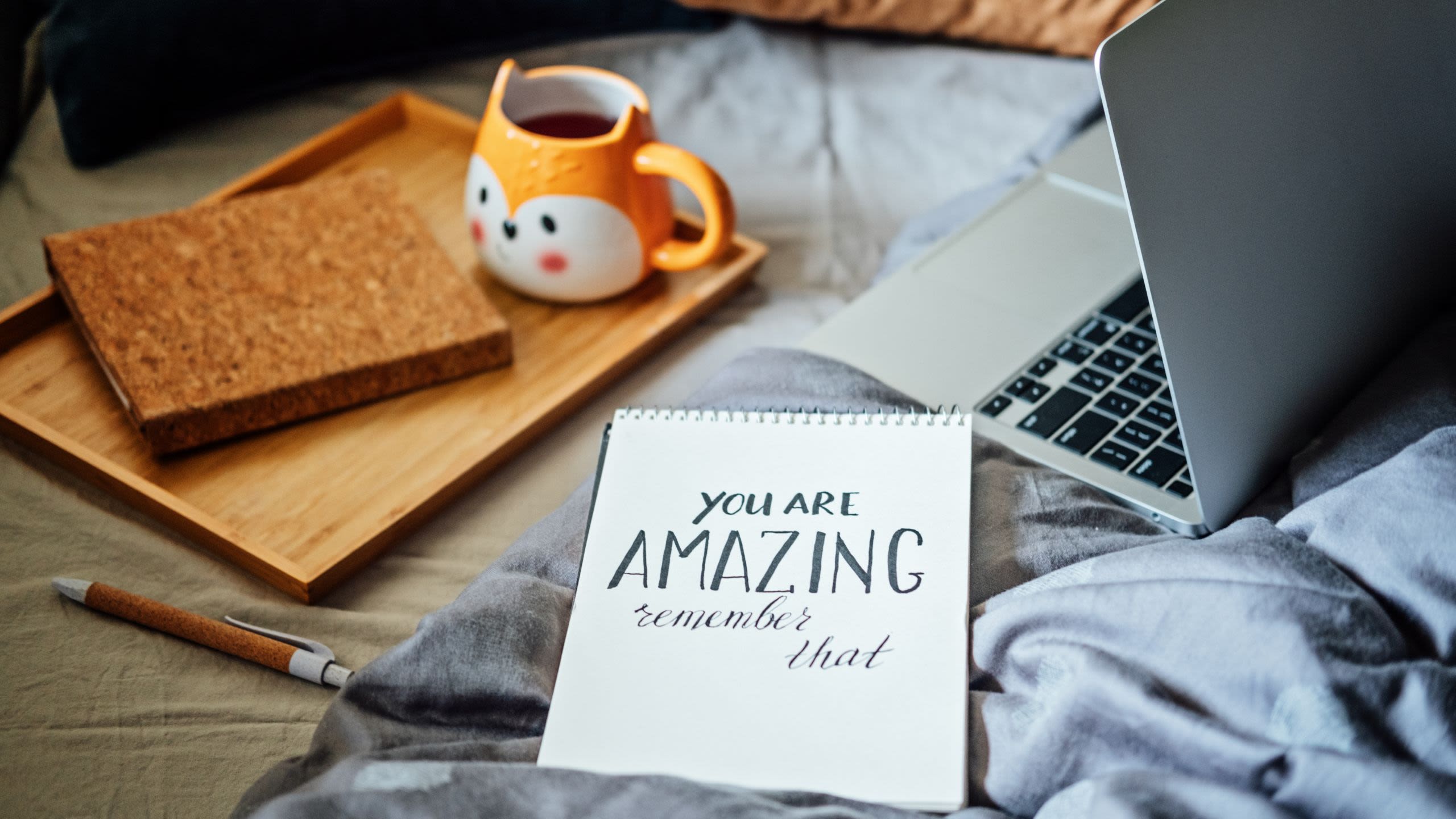 A notepad reads: "You are amazing remember that" with a laptop, notepad, pen and mug on a tray behind