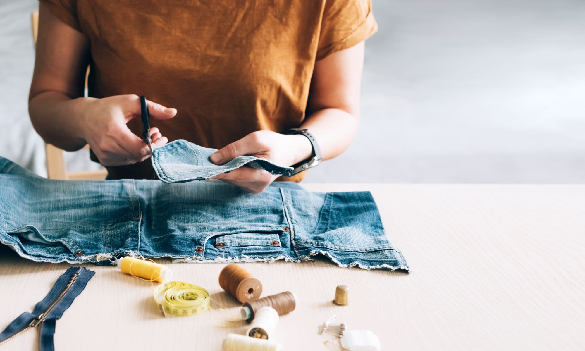A woman cuts a pocket from a pair of jeans with thread and a tape measure on the table