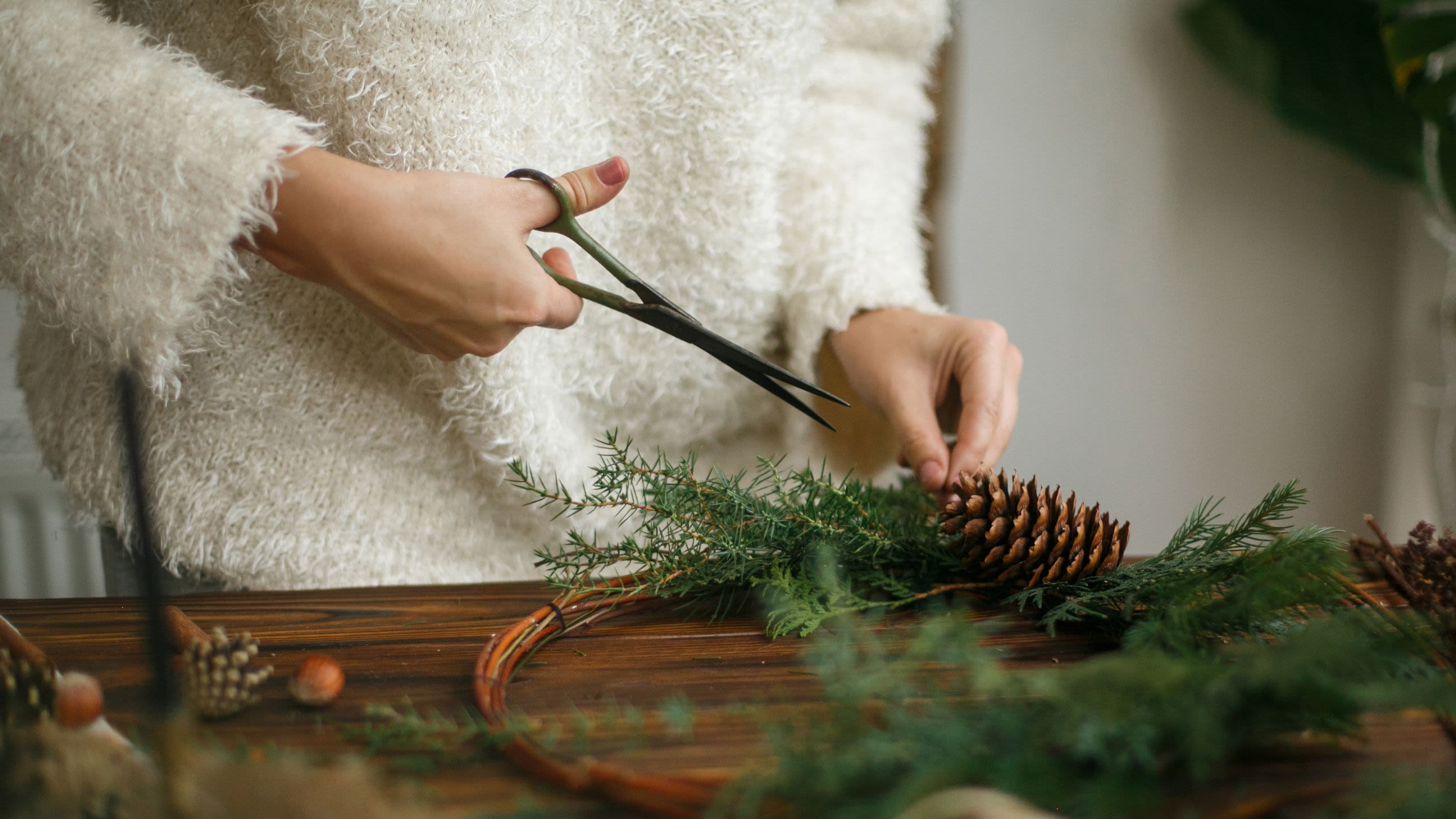 A person (face not in shot) crafts a festive wreath from natural materials including a pine cone