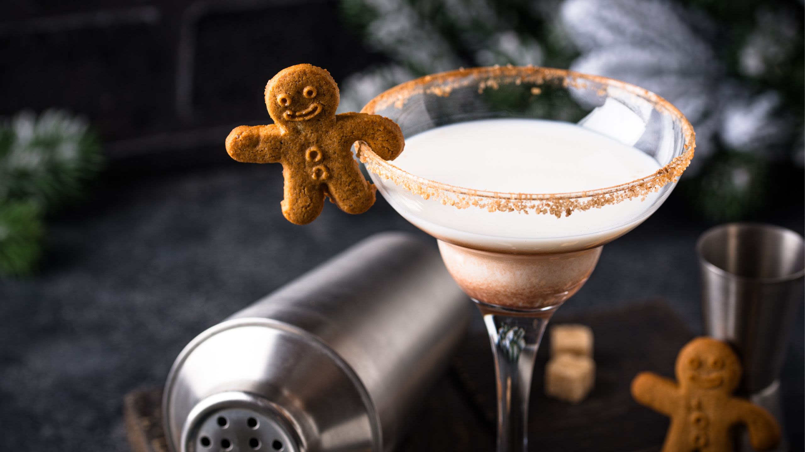 A cocktail shaker lies on the counter with a cocktail glass filled with a festive drink and a gingerbread garnish